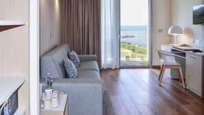 Quarto moderno com sofá cinzento, secretária de madeira e vista para o mar através de uma porta de vidro.