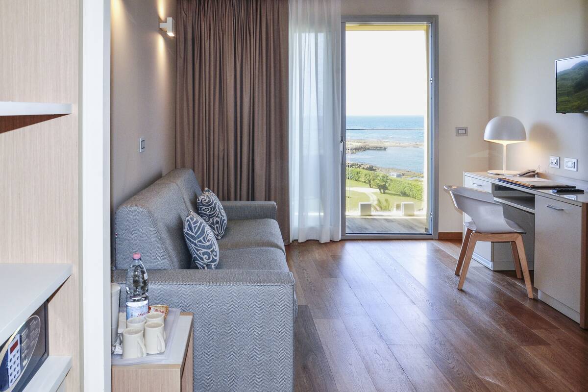 Une chambre moderne avec un canapé gris, un bureau en bois et une vue sur la mer, visible à travers une porte vitrée.