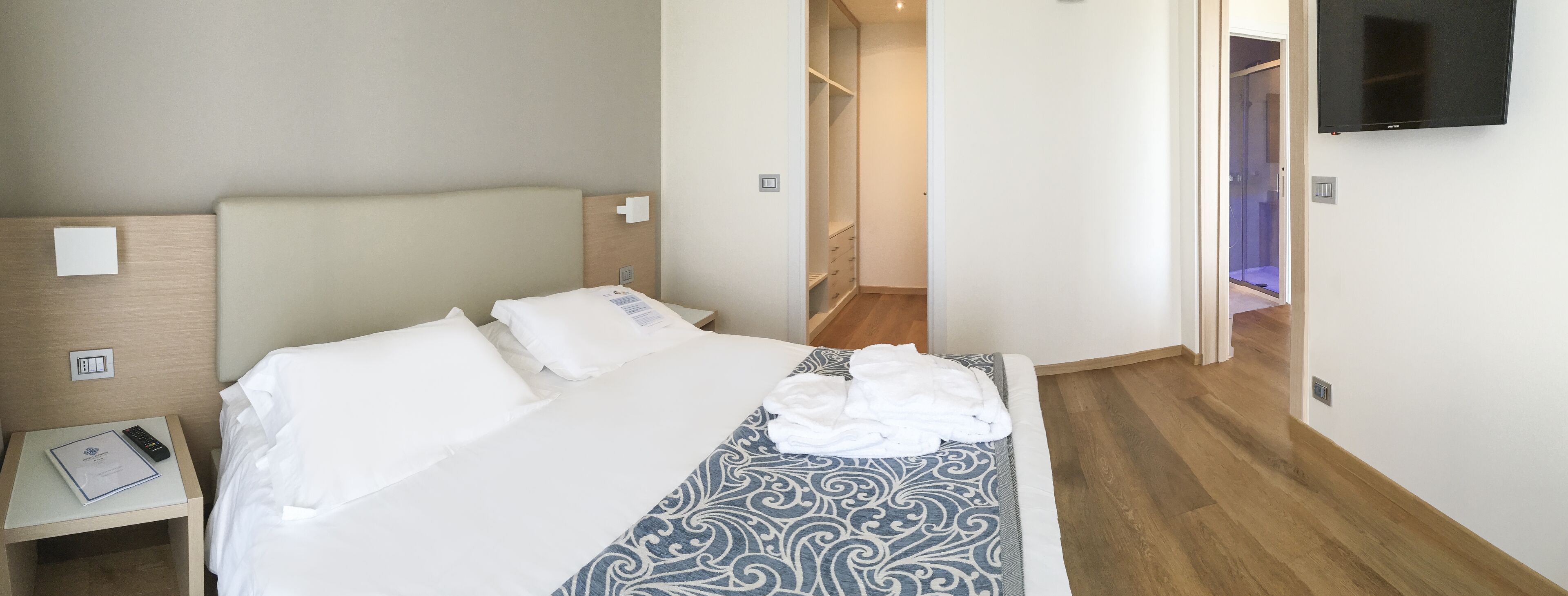 A hotel room with a bed, a bedside table with a remote control and a booklet, a patterned throw blanket, and a TV mounted on the wall.