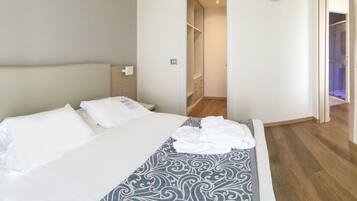A hotel room with a bed, a bedside table with a remote control and a booklet, a patterned throw blanket, and a TV mounted on the wall.