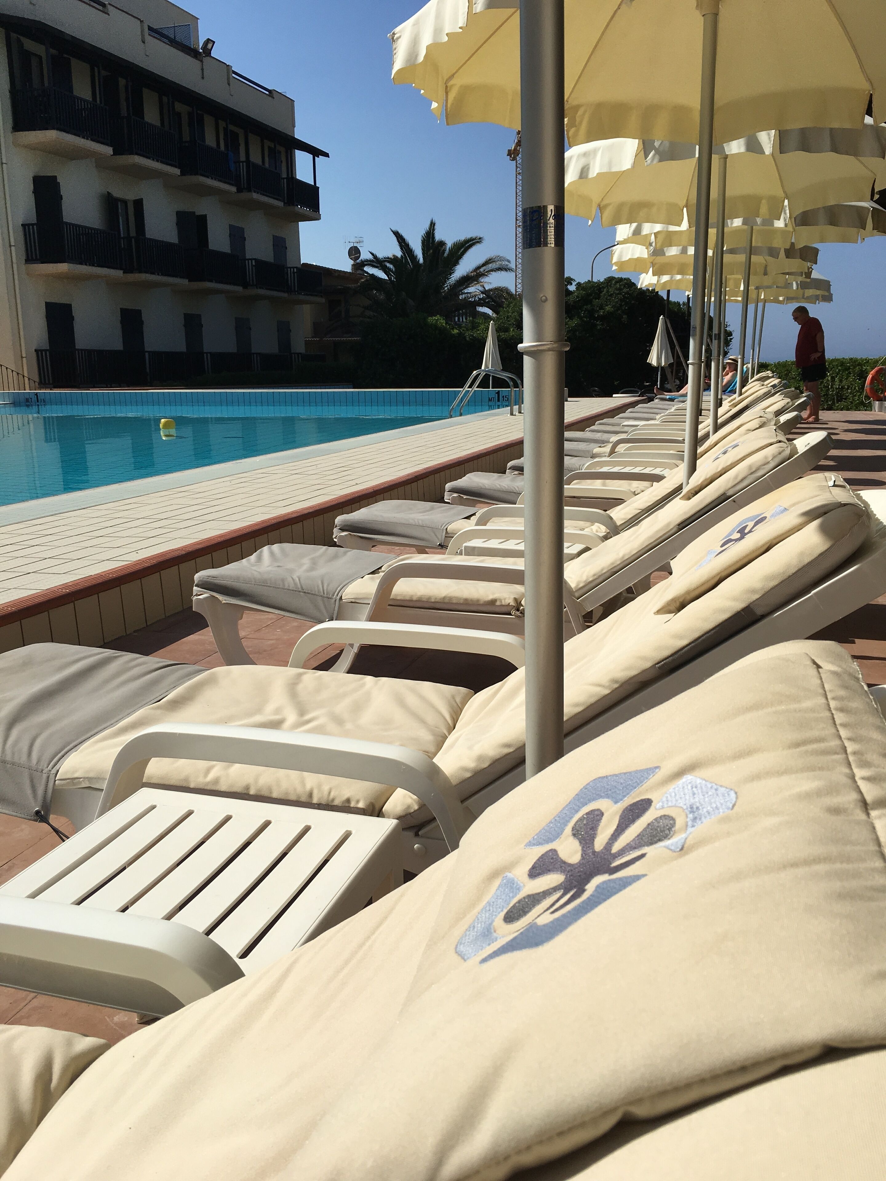 Zona de piscina con sillones, sombrillas, una piscina y un edificio al fondo.