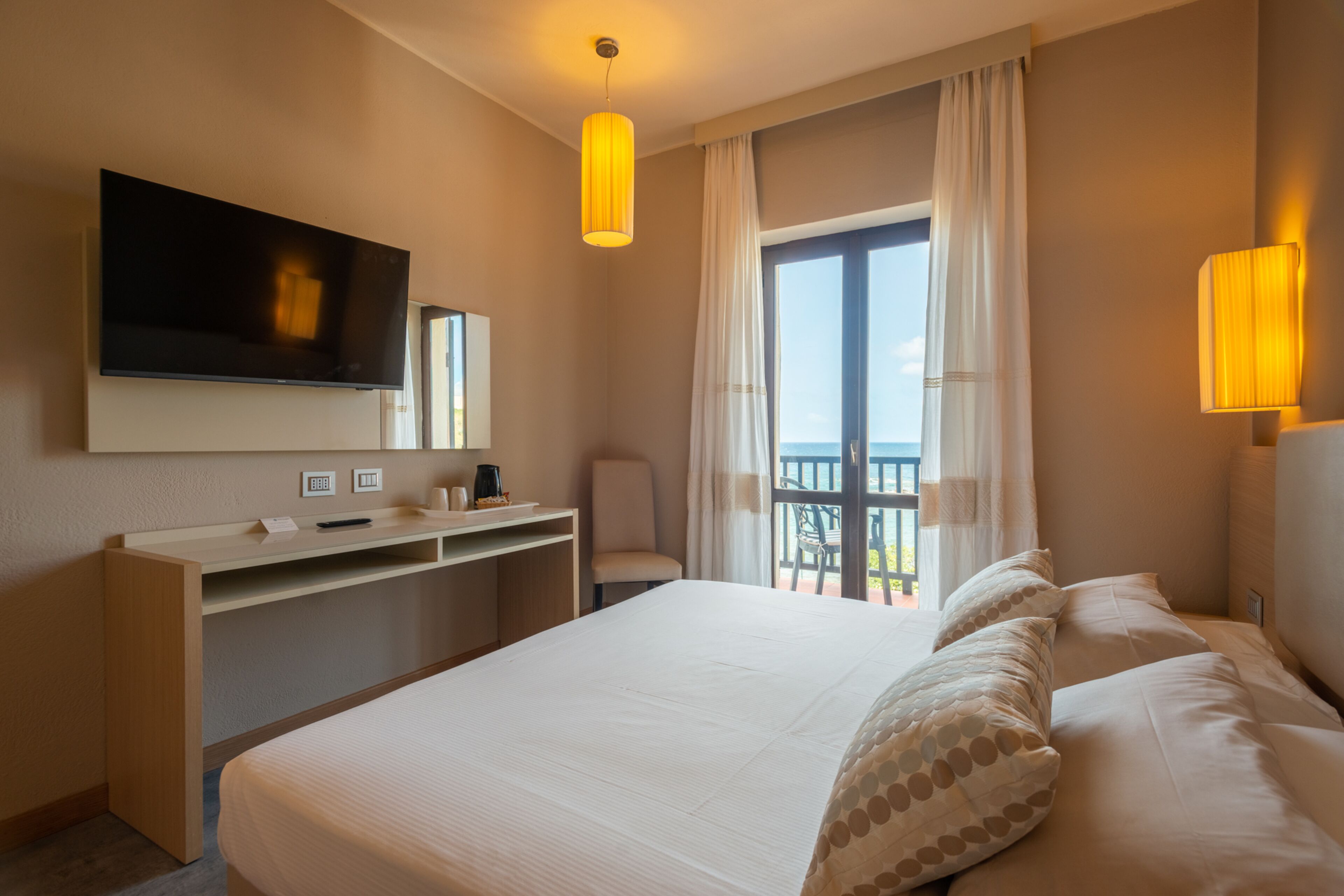 A hotel room with a large bed, a desk with a flat-screen TV, a chair, and a balcony with curtains.