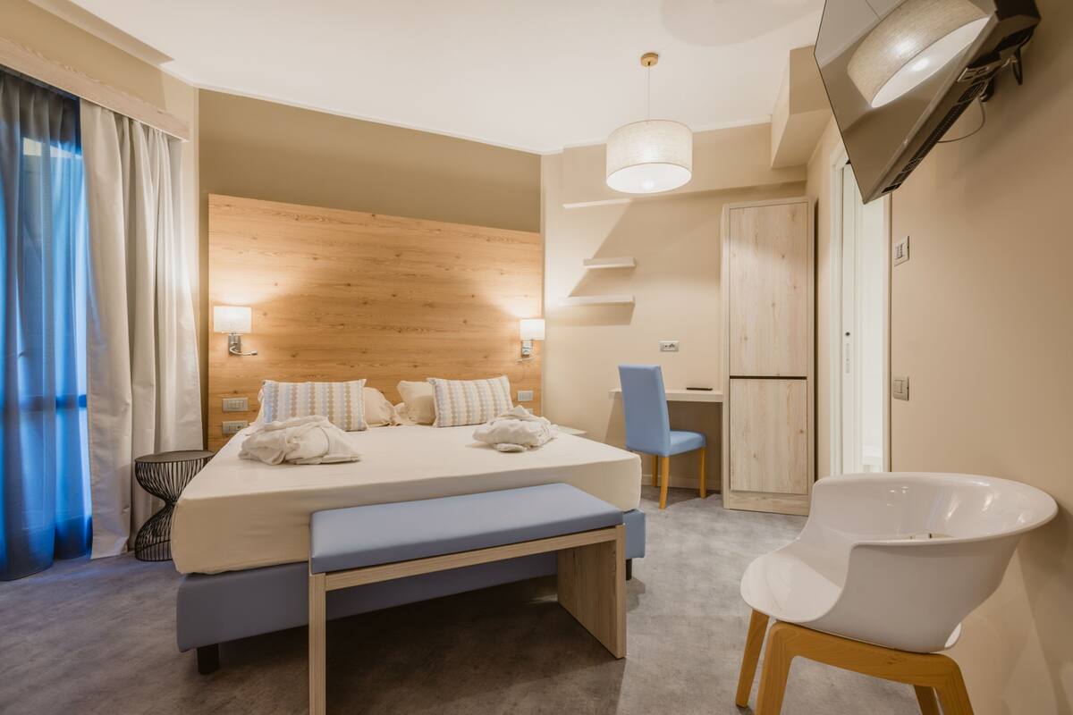 Une chambre d’hôtel moderne avec une tête de lit en bois, un grand lit avec une banquette, un bureau, une chaise et un téléviseur fixé au mur.