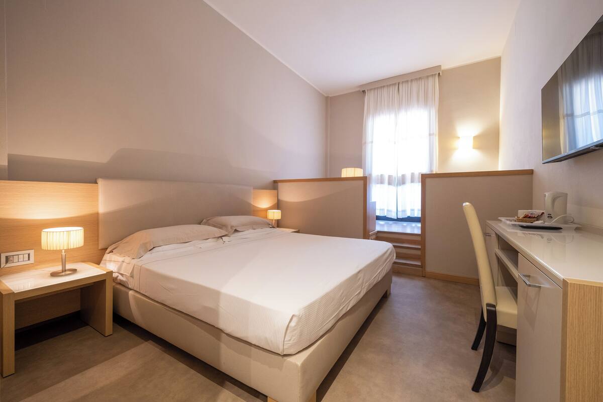 Une chambre d’hôtel équipée d’un lit, d’une table de chevet, d’une lampe, d’un bureau, d’une chaise et d’un téléviseur fixé au mur.