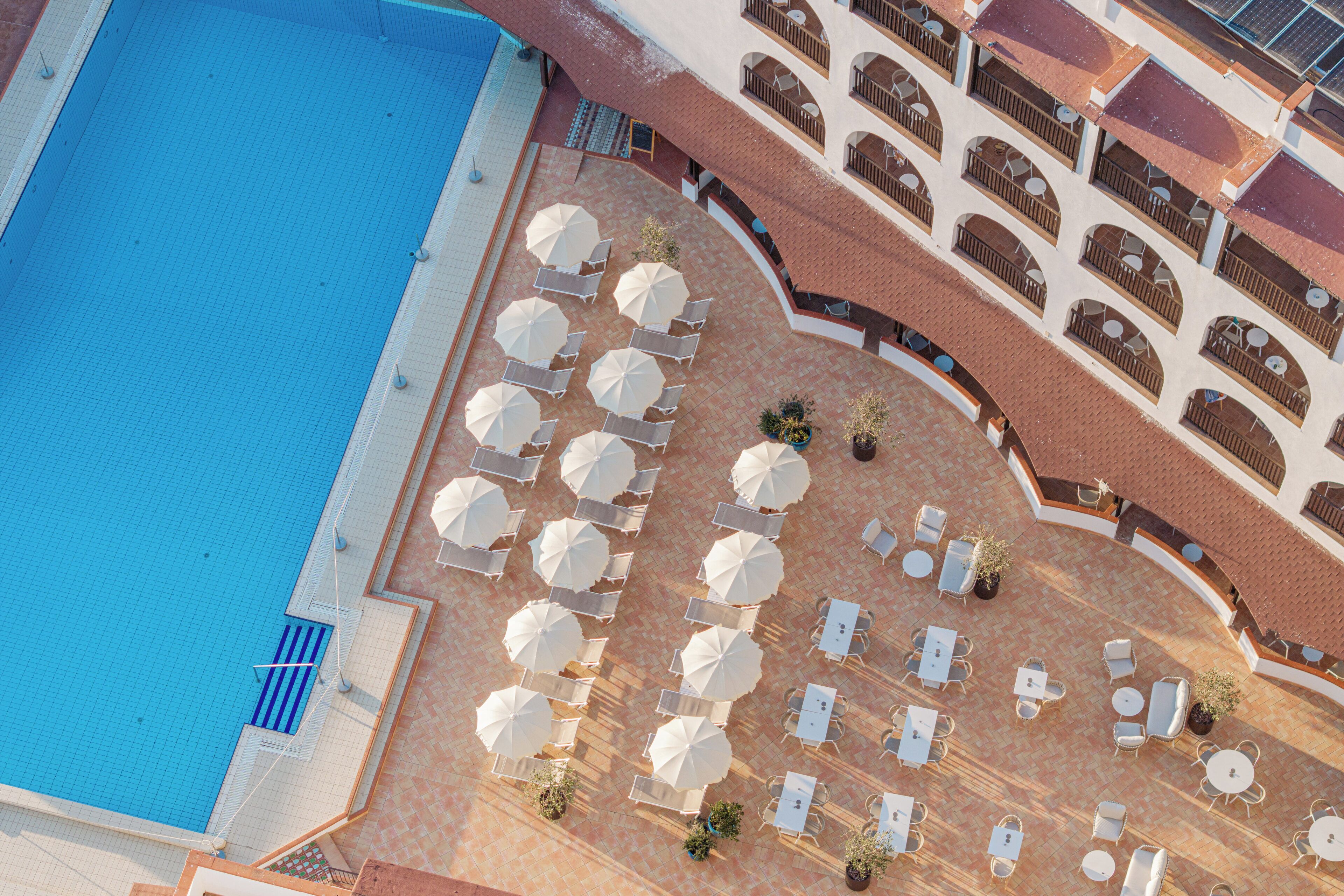 An aerial view of a hotel with a swimming pool, outdoor seating area with umbrellas, and a covered patio with tables and chairs.