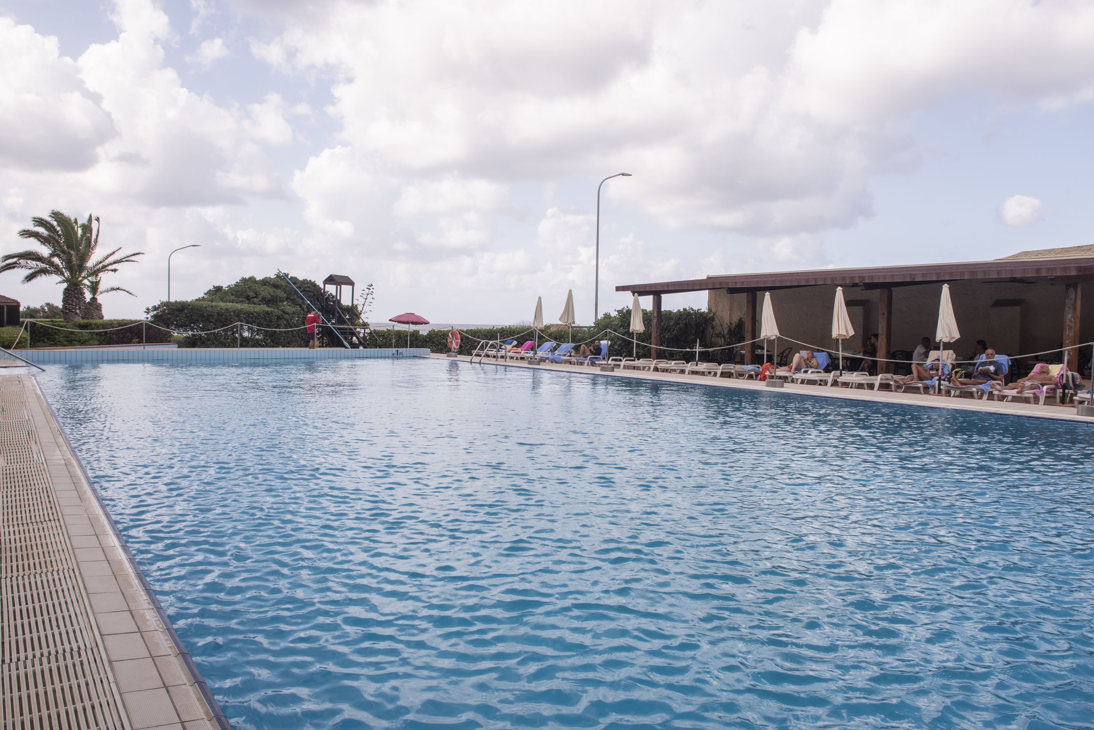 A swimming pool with lounge chairs and umbrellas, surrounded by a covered area with a palm tree and a slide in the background.