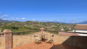 Una terrazza con un muro in pietra, un tavolo e due sedie in vimini. La terrazza si affaccia su un paesaggio collinare con case sparse e cielo sereno.