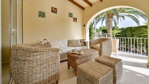 A cozy lounge area with wicker furniture, a white sofa, and a balcony with a view of palm trees. - Holiday Villa Moraira with private heated pool in walking distance to beaches (Moraira)
