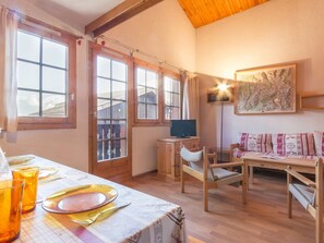 Ein gemütliches Zimmer mit Holztisch, gedecktem Esstisch, einer Couch, einem Fernseher und einem großen Fenster mit Vorhängen.