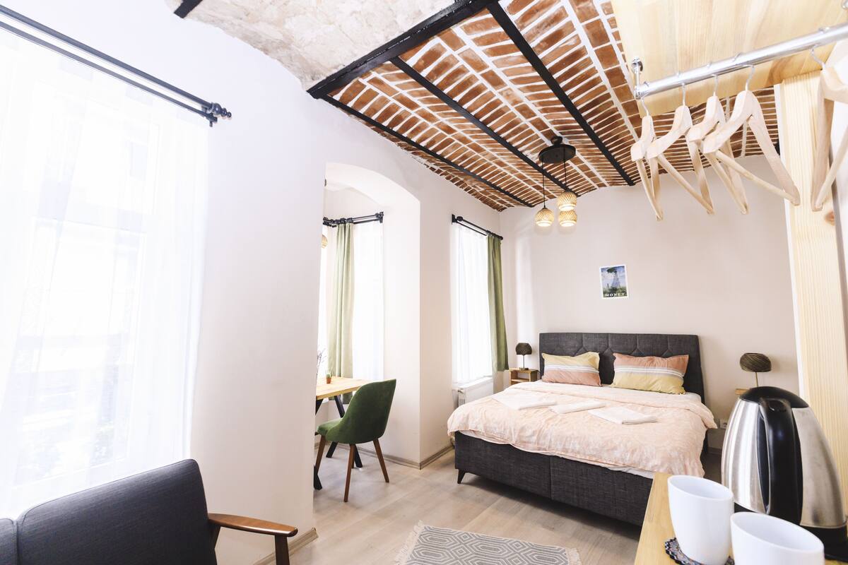 A modern bedroom with a bed, a desk, and a chair. There is a window with curtains, a radiator, and a hanging light fixture.
