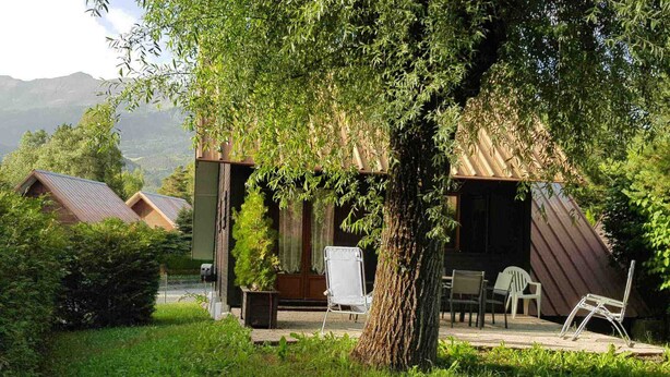 Un chalet rustique doté d’une terrasse en bois, entouré d’une végétation luxuriante et avec une chaîne de montagnes en toile de fond.