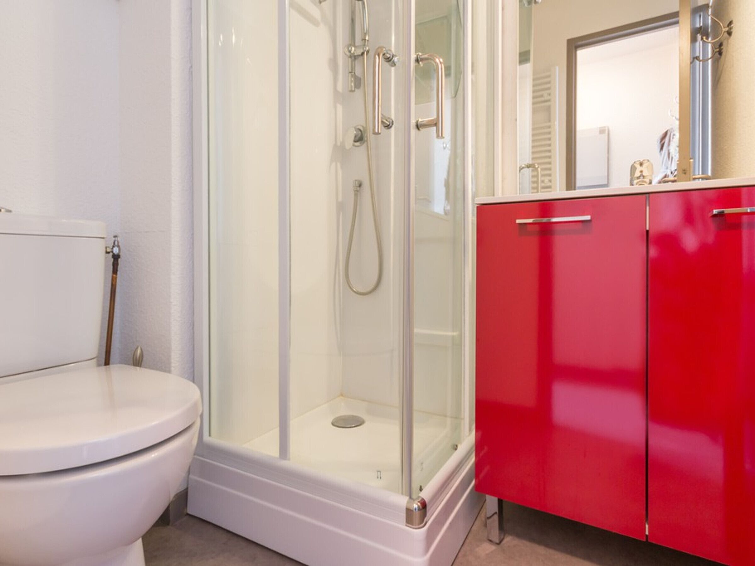Une salle de bain avec une douche à parois de verre, des toilettes blanches et un meuble rouge.