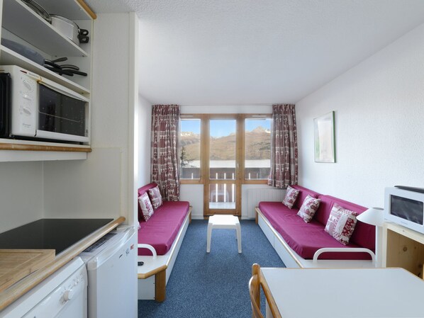 A room with a bed, a table, and a chair. There is a window with curtains, a microwave, and a refrigerator.