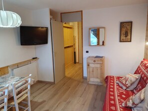 A compact living space with a television mounted on the wall, a wooden cabinet, a red patterned sofa, and a dining area with a table and chairs.