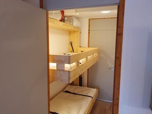 A small, narrow room with a bunk bed and a single bed. There is a wooden shelf above the bunk bed with various items on it.