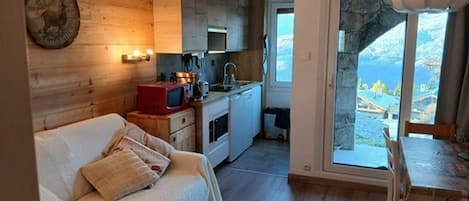 A cozy cabin with a wooden interior, a kitchenette, a sofa with cushions, and a view of the mountains through a large window.