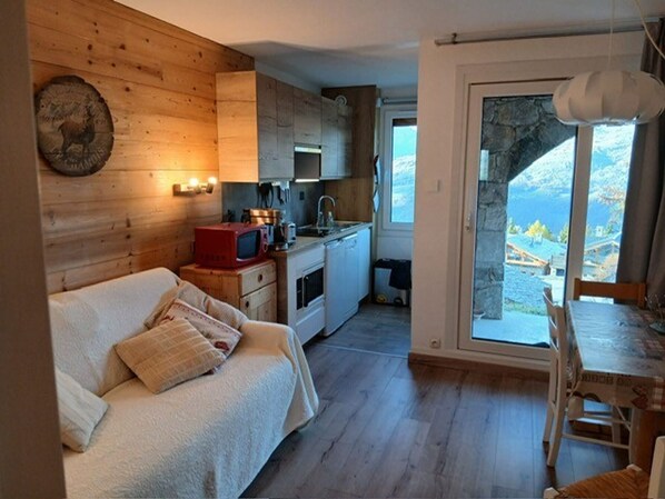 A cozy cabin with a wooden interior, a kitchenette, a sofa with cushions, and a view of the mountains through a large window.
