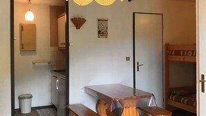 A compact room with a kitchenette, a wooden table with benches, a bed, and a ceiling light fixture.