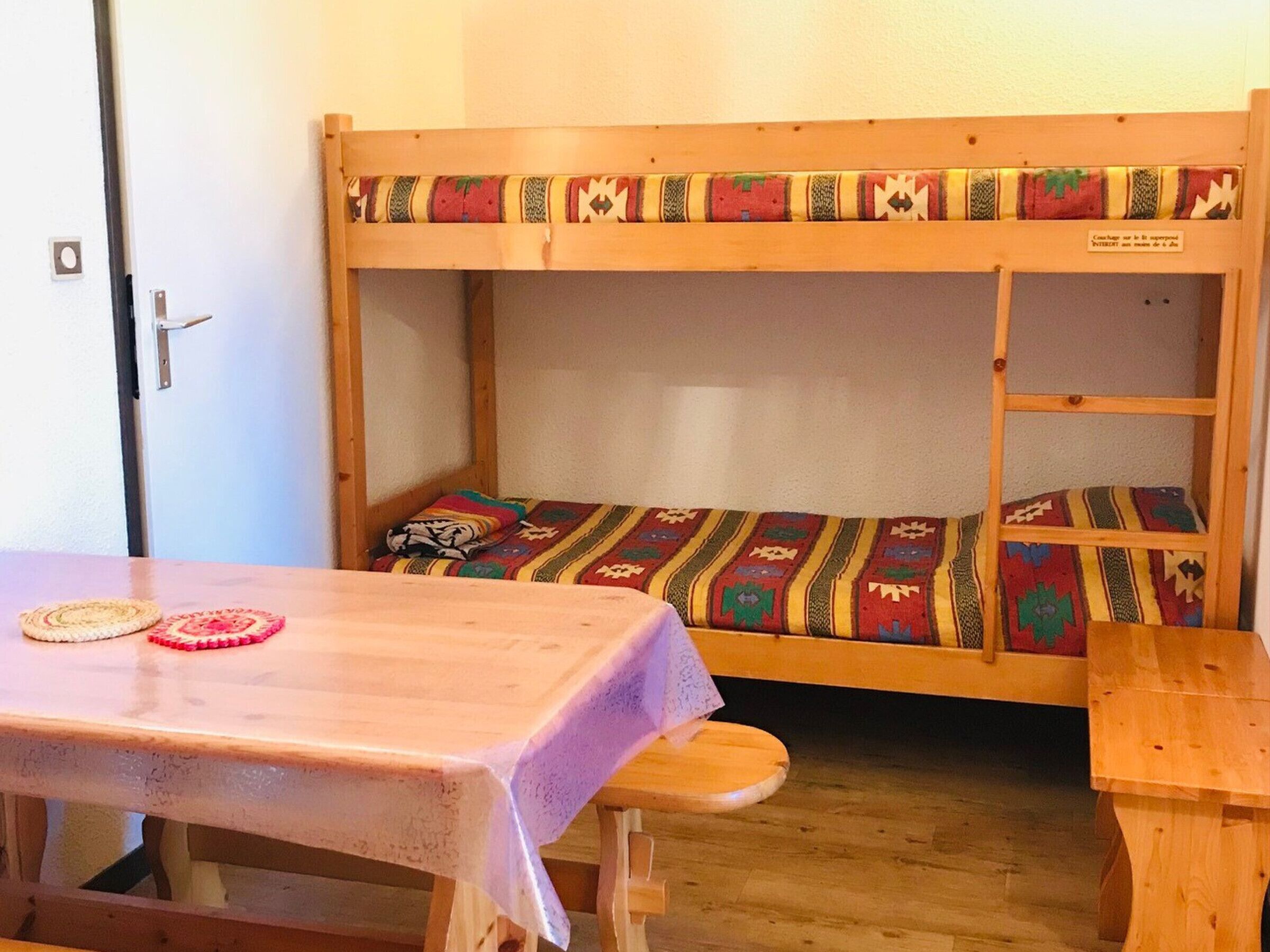 Un lit superposé avec une couverture à motifs, une table en bois recouverte d’une nappe et un banc.