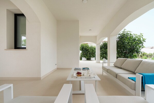 A modern, minimalist living area with a white sofa, a coffee table with a tray of fruit and cups, and a view of greenery outside.
