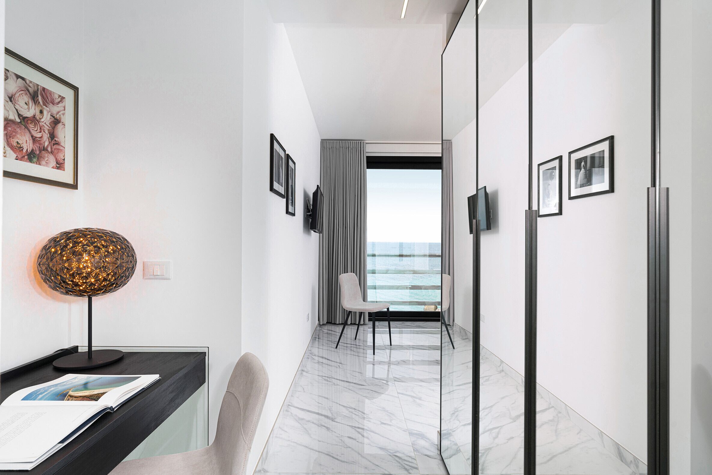 Une chambre d’hôtel moderne avec un sol en marbre, un bureau avec une lampe, une chaise et une vue sur la mer par une grande fenêtre.