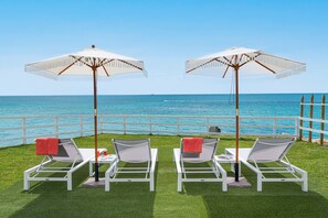 Two white lounge chairs with red towels, each under a white umbrella, on a grassy area with a view of the sea.