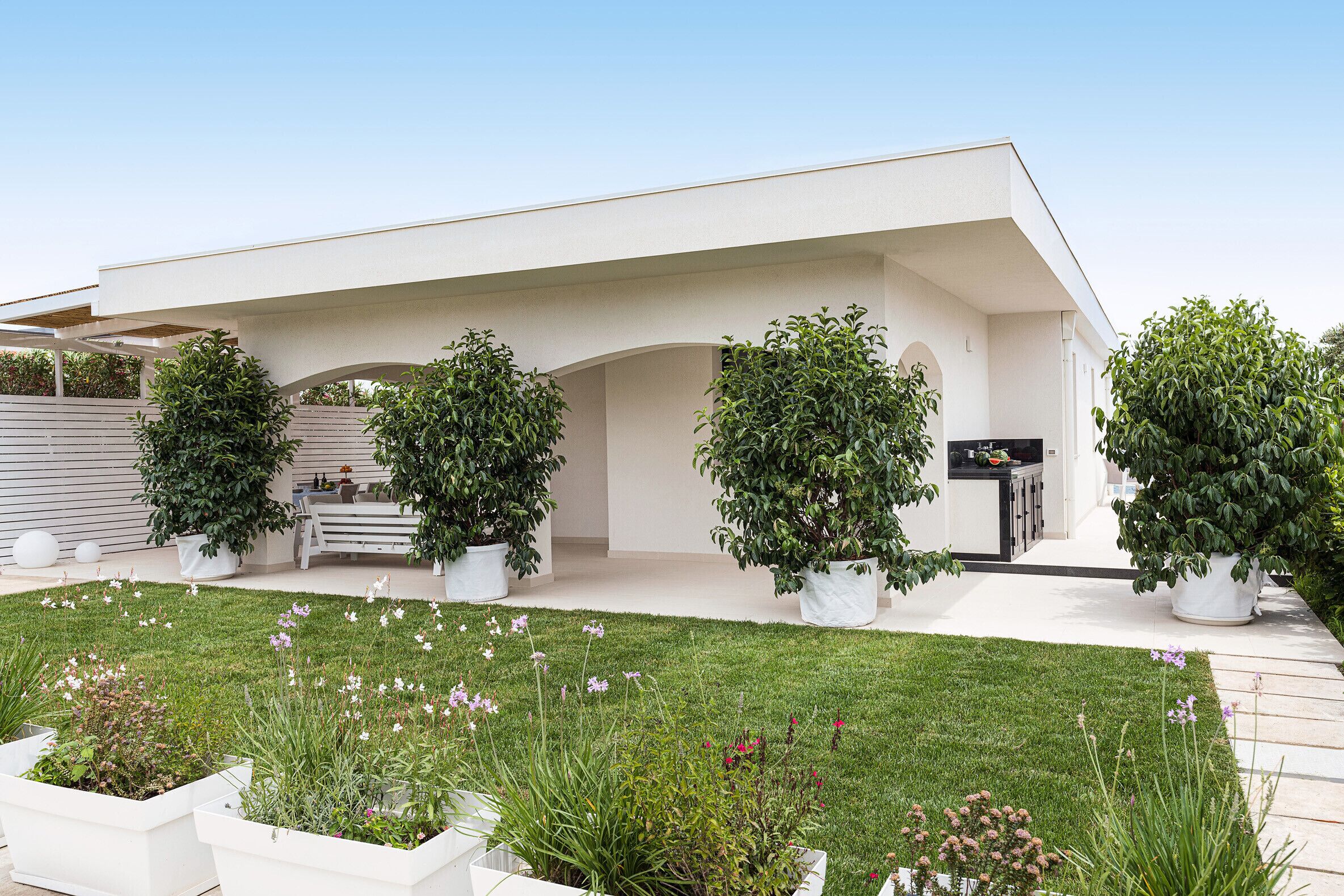 A modern building with a flat roof, white exterior walls, and a covered outdoor area with potted plants and a grassy lawn.