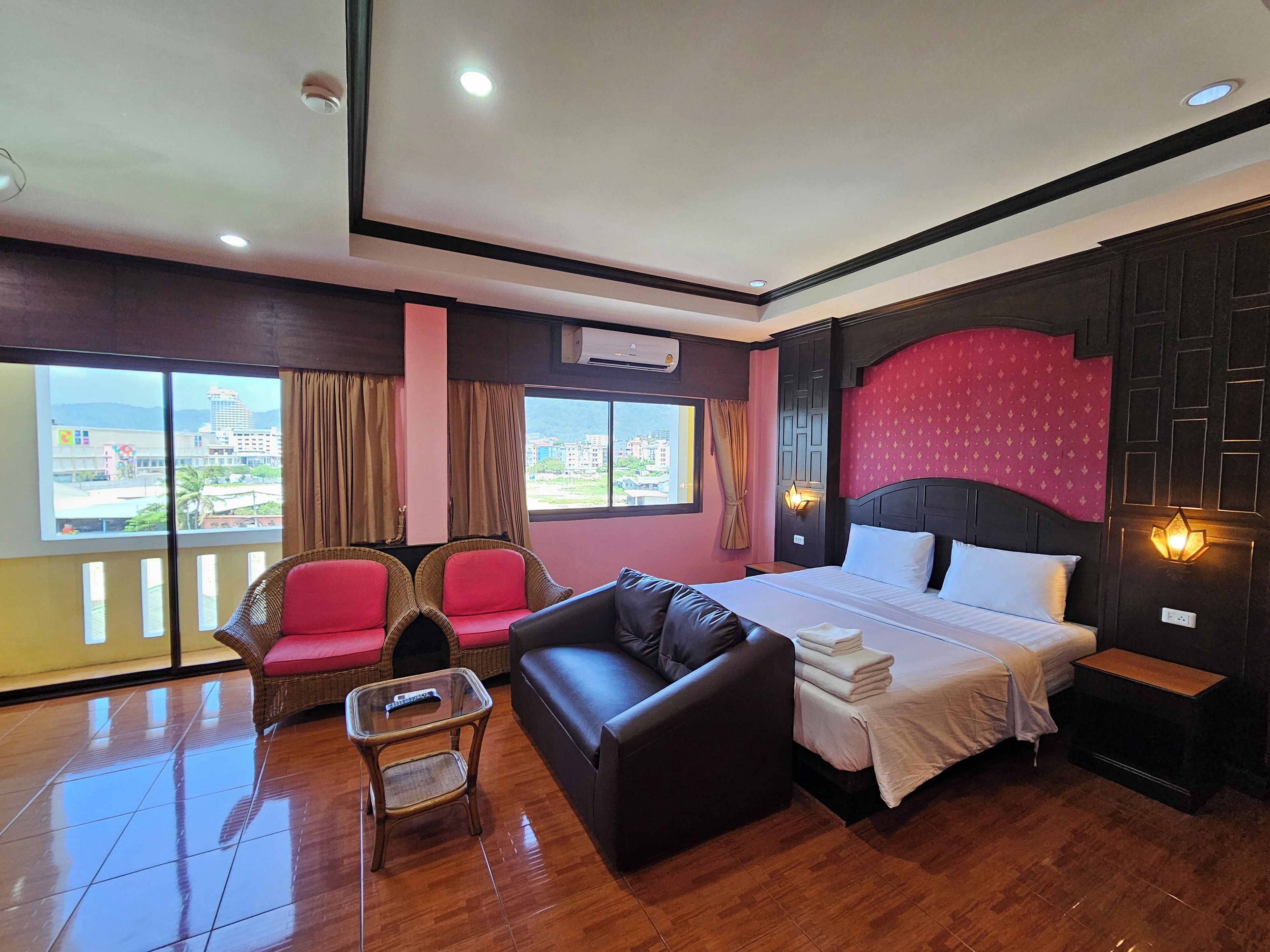 Quarto de hotel com uma cama grande, um sofá, duas poltronas, uma mesinha e vista para a cidade através de grandes janelas.