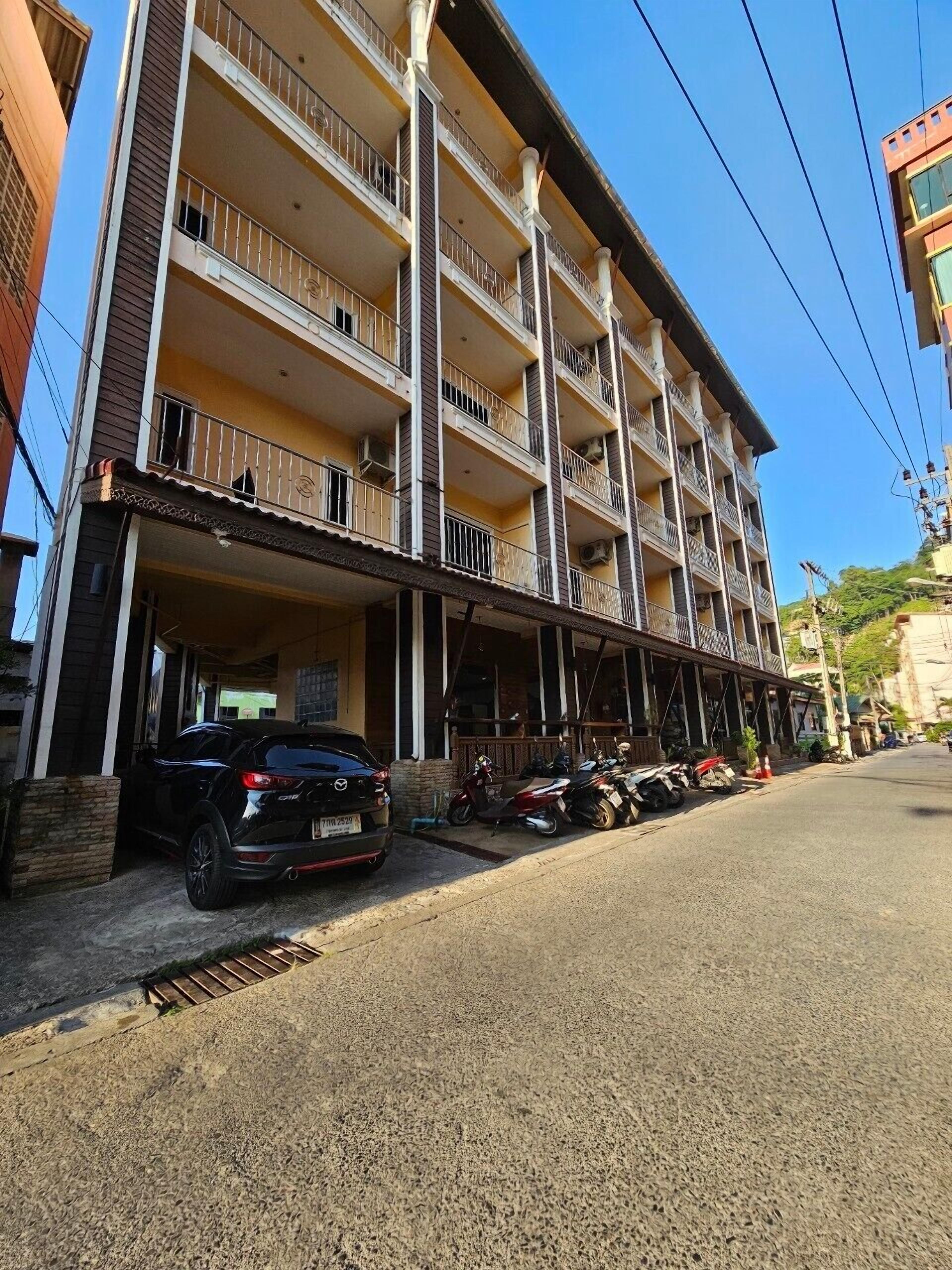 Un edificio de varios pisos con balcones, un acceso cubierto y un estacionamiento con varias motocicletas y un automóvil.