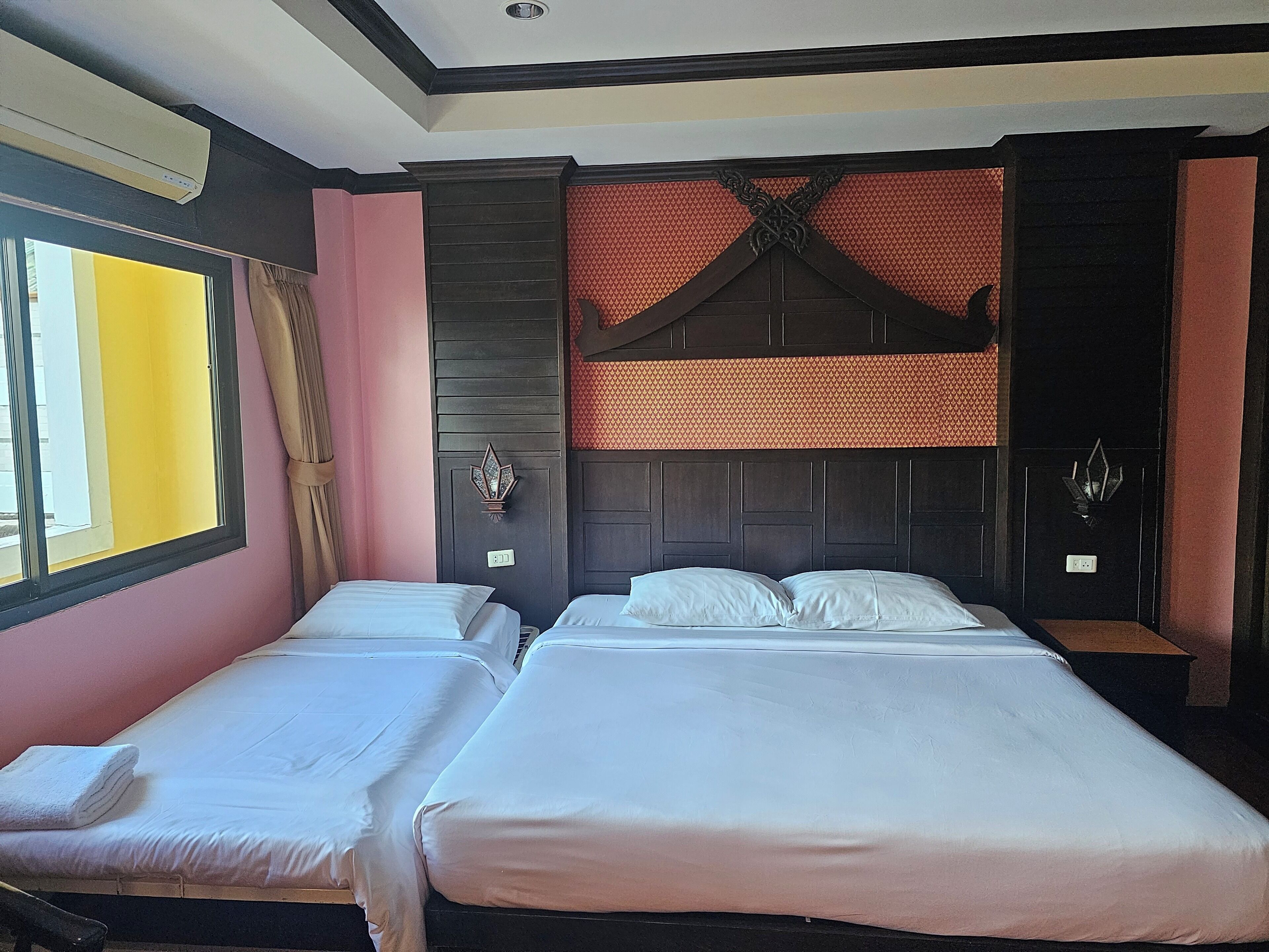 A hotel room with two single beds, a window with curtains, a decorative wall panel, and a ceiling vent.