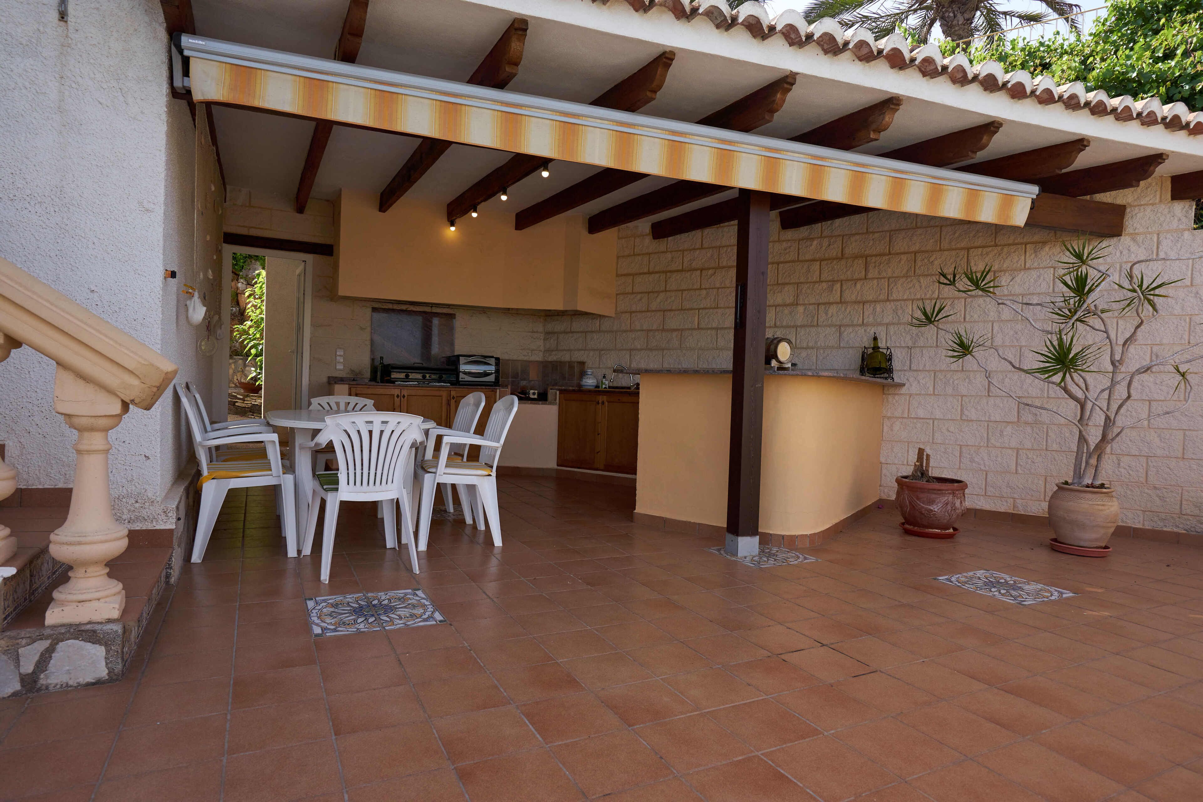 A covered patio with a dining area, kitchen, and potted plants.