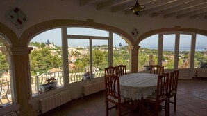 A dining area with a table and chairs, large arched windows, and a view of a coastal town. - Luxurious 3 bedroom villa with stunning panoramic views in Denia (Denia)