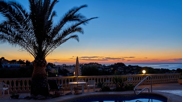A pool area with a palm tree, lounge chairs, and a view of the sunset over the ocean. - Luxurious 3 bedroom villa with stunning panoramic views in Denia (Denia)
