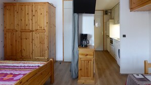 Una habitación pequeña y funcional con cama, un armario de madera, un escritorio pequeño con cafetera y una kitchenette con refrigerador y lavadora.