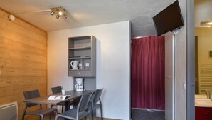 A compact room with a wooden wall, a table with chairs, a coffee machine, a TV mounted on the wall, and a red curtain. - Renovated and Comfortable Studio · Heart of Resort (Plagne Bellecôte)