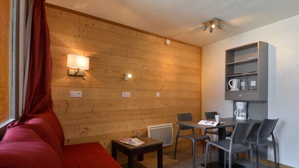 A cozy room with a wooden wall, a red sofa, a small table with chairs, and a coffee machine on a shelf. - Renovated and Comfortable Studio · Heart of Resort (Plagne Bellecôte)