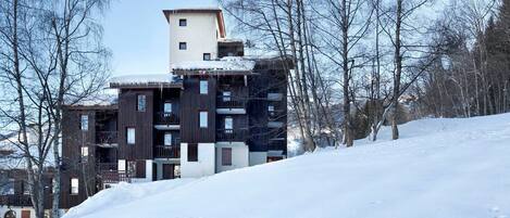 A modern multi-story building with a mix of dark and light facades, surrounded by snow-covered ground and leafless trees.