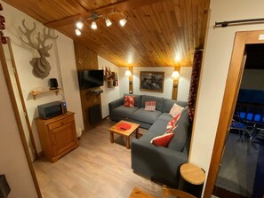 Un accogliente soggiorno con soffitto in legno, un divano grigio con cuscini rossi e bianchi, una televisione e un piccolo tavolino in legno.