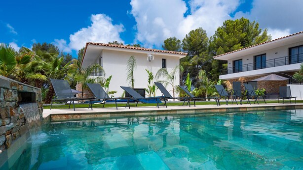 A swimming pool with lounge chairs, a stone water feature, and a modern house with a tiled roof.