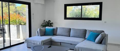 A modern living room with a grey sectional sofa, a round coffee table, and a large sliding glass door leading to an outdoor area with plants.