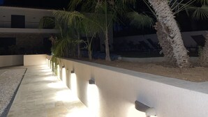 A nighttime outdoor area with a pathway, palm trees, and a modern building with a flat roof and large windows.
