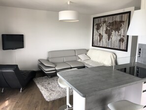 En moderne opholdsstue med en grå sofa, et lille spisebord, en sort stol, en hvid måtte og et ophængt fjernsyn.