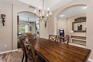 A dining area with a wooden table and chairs, a chandelier, and a kitchen in the background. - Serenity Now by Shine Villas at Watersong #416 (Davenport)