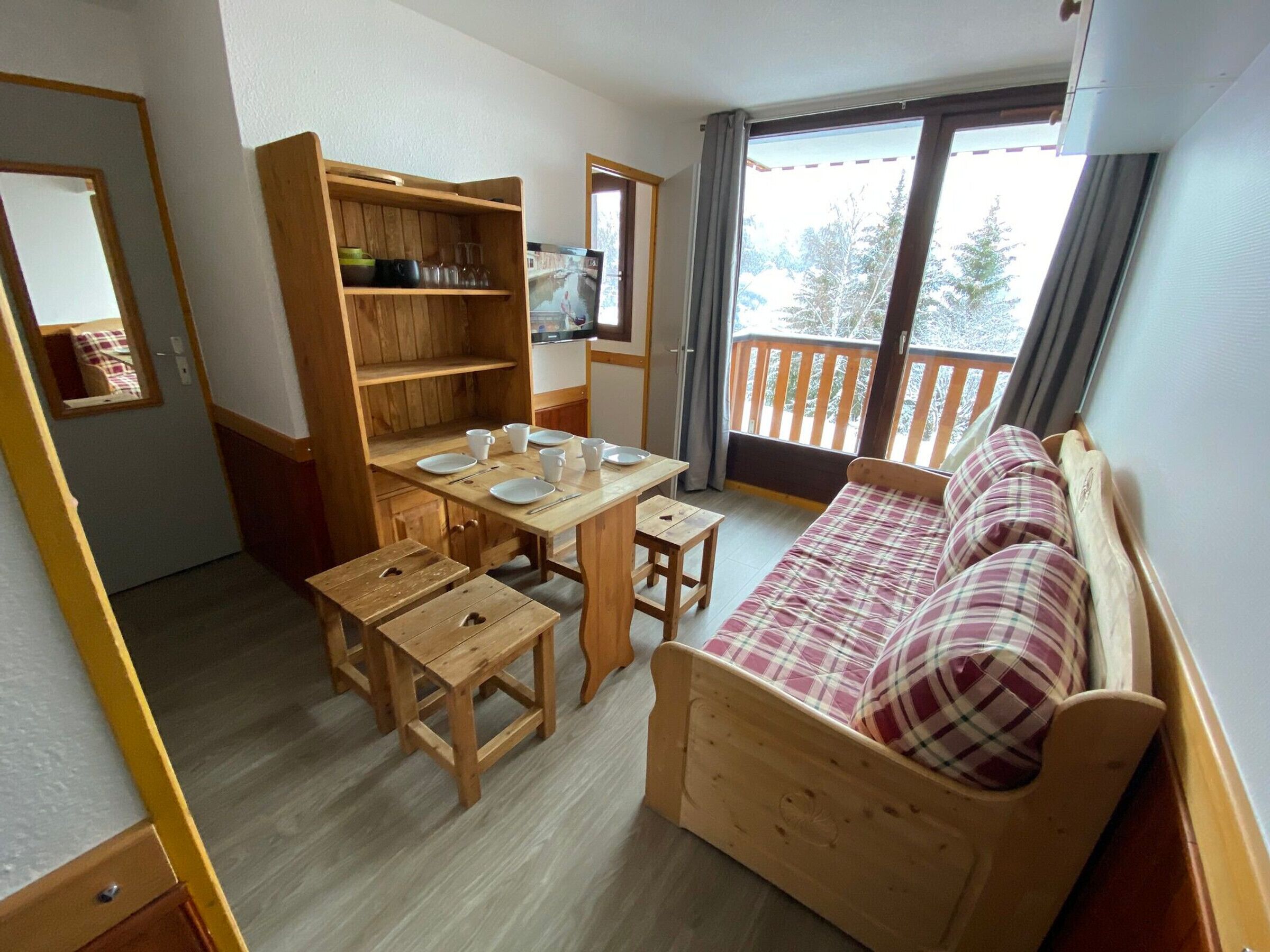 Una habitación acogedora con una mesa de madera, dos sillas, un pequeño armario de madera, un sofá a cuadros y un balcón con vistas a árboles.