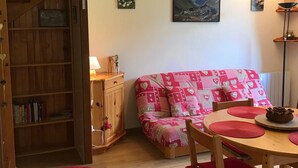 A cozy room with a wooden bookshelf, a red sofa with patterned cushions, a wooden dining table with a teapot, and a mountain landscape painting on the wall.