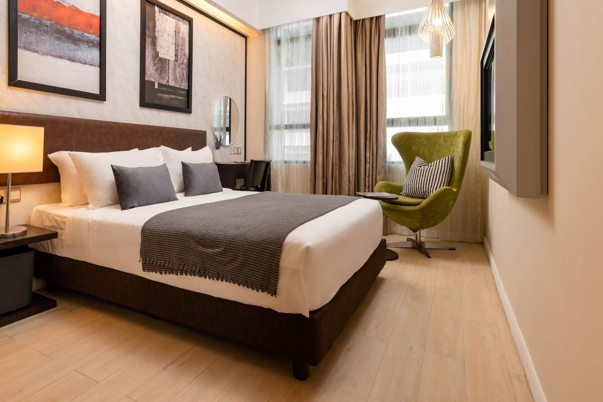 A neatly arranged hotel room with a large bed, a bedside table with a lamp, a chair, and a window with curtains.