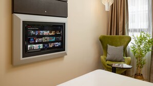 A hotel room with a mounted TV displaying various TV shows, a green armchair with a patterned pillow, a small table with a remote control, and a potted plant. - Centrestage Petaling Jaya- Perfect Host (Petaling Jaya)