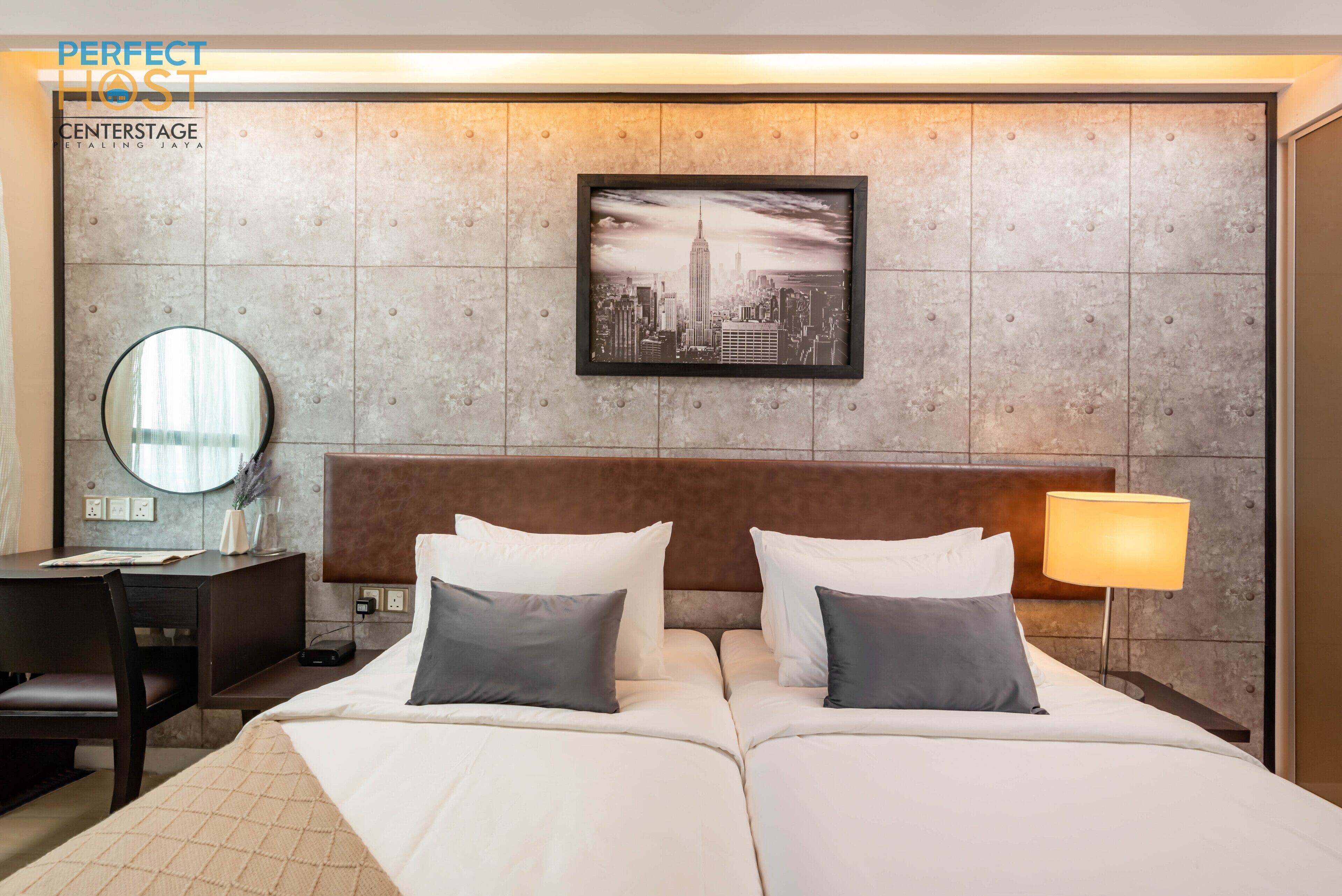 A hotel room with a large bed, a desk, a chair, a round mirror, a lamp, and a framed picture of a cityscape.