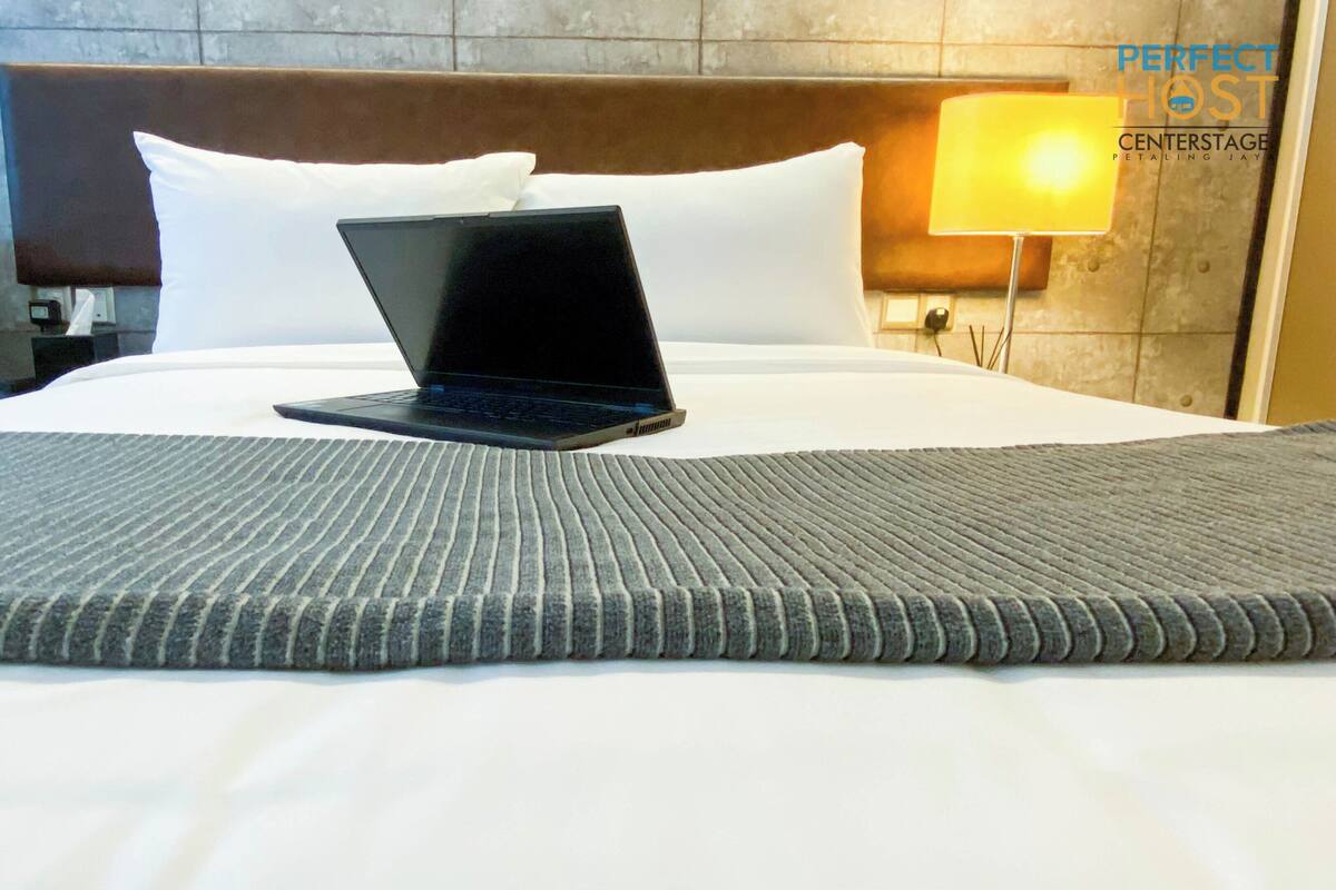 A hotel room with a bed, a laptop on the bed, a lamp, and a nightstand with a bottle and glasses.