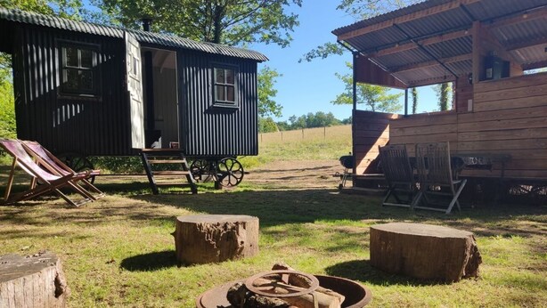 Una cabaña rústica de madera, con un pequeño porche y sillas, y un carretón con leña al frente.